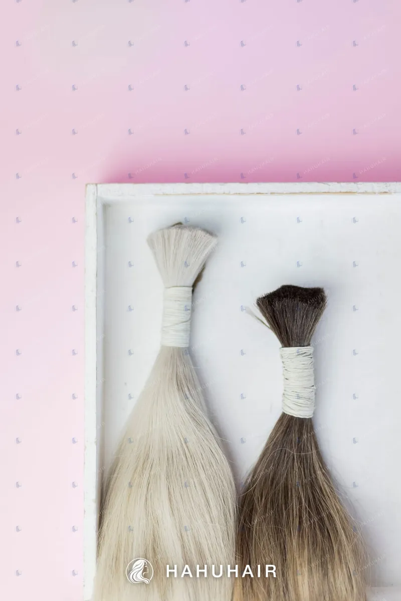 Two tied bundles of bulk human hair, one ash blonde and one brown, in a white tray