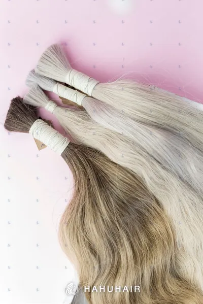 Several tied bundles of bulk human hair in ash blonde and brown shades on a pink background